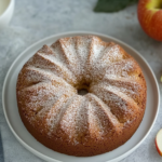 Apple Cider Donut Cake topped with cinnamon sugar, served on a plate with a slice removed to show the fluffy texture