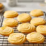 A stack of golden-brown, round crispy butter cookies on a white plate, with one cookie broken in half to show its delicate, snappy texture.