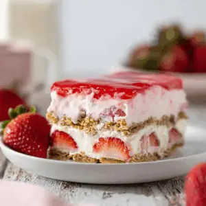 A perfect slice of no-bake Strawberry Eclair Cake on a white plate. The visible layers include graham crackers, creamy cheesecake filling, sliced strawberries, and a sweet pink frosting on top.